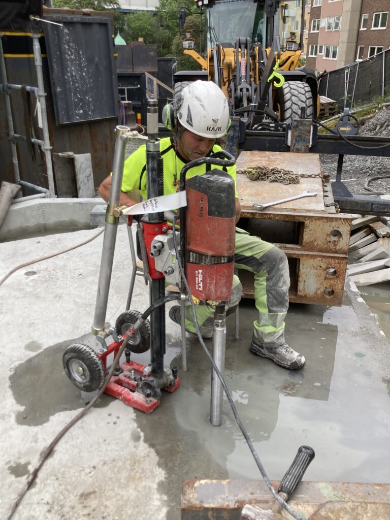 Tekniker använder röd kärnborrmaskin på stativ för borrning i betongplatta på urban byggarbetsplats