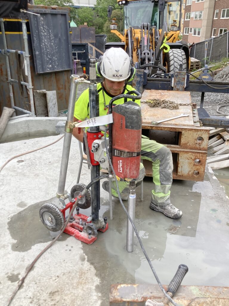 Kärnborrning i betong med Hilti borrmaskin på stativ, tekniker i hjälm och varselkläder på byggarbetsplats