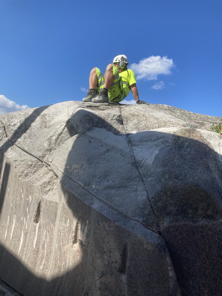 Tekniker i varselkläder och hjälm sitter på toppen av stort bergblock med synliga skärspår i berget