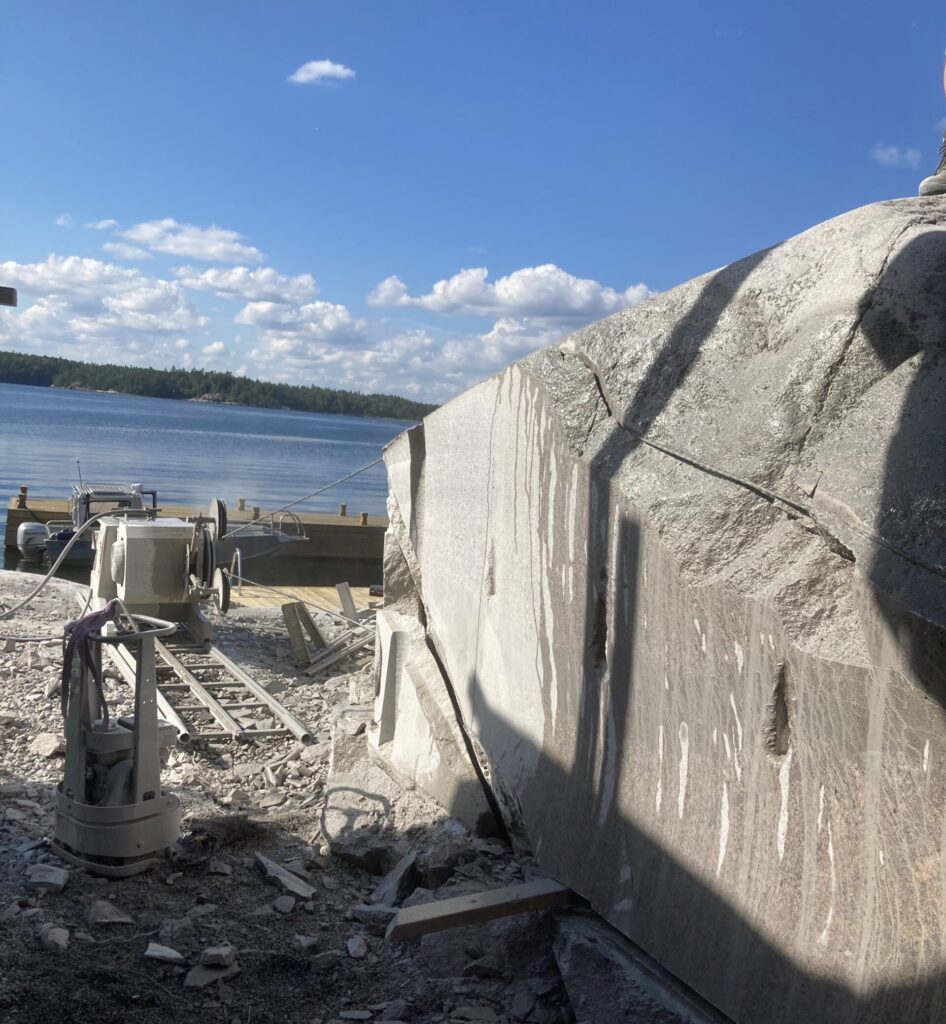 Vajersågsmaskin på räls vid strandkant med stort bergblock under pågående vajersågning