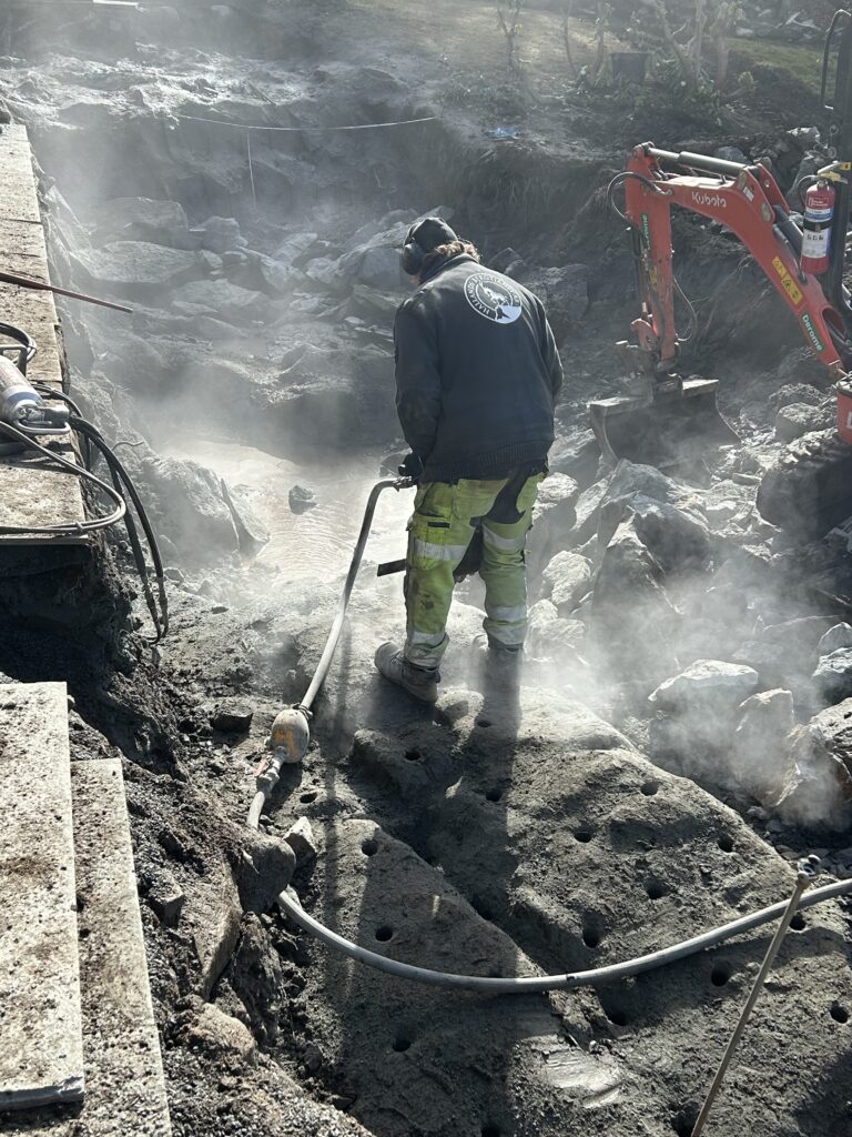 Arbetare utför hydraulspräckning av berg på arbetsplats i Halland med synlig ånga och minigrävare i bakgrunden